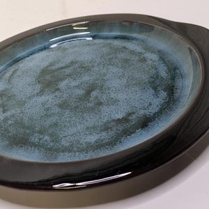 以下が含まれることがあります： ダークで光沢のある縁と取っ手が付いた、楕円形のセラミック製サービング皿。内側はまだらな青緑色の釉薬が特徴です。この皿は料理を盛り付けるように設計されており、テーブルセッティングにエレガントさを加えます。