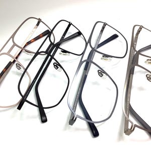 May include: Four pairs of eyeglasses with various frame colors, including a light brown tortoiseshell, black, and gray. The glasses have a modern, geometric design with clear lenses and nose pads. The frames are made of metal.