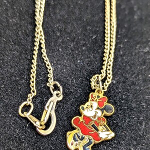 May include: Gold-toned necklace featuring a Minnie Mouse pendant. The pendant is a colorful enamel design of Minnie Mouse in a red dress, black skirt, and yellow shoes. The necklace has a delicate chain and a clasp.