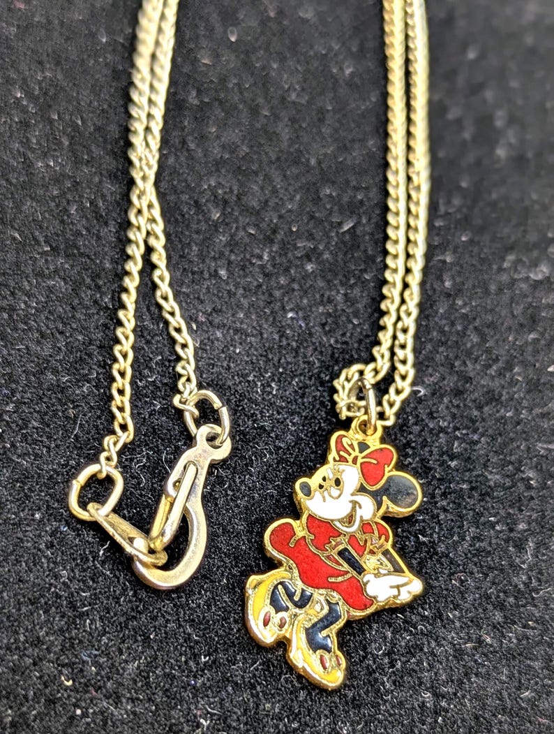 May include: Gold-toned necklace featuring a Minnie Mouse pendant. The pendant is a colorful enamel design of Minnie Mouse in a red dress, black skirt, and yellow shoes. The necklace has a delicate chain and a clasp.
