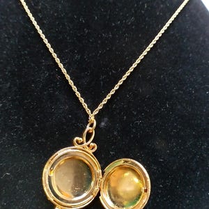 May include: A gold-colored locket necklace with an open, round pendant. The pendant is hinged and opens to reveal a small space. The necklace chain is a delicate, twisted design. The locket is displayed against a black background.