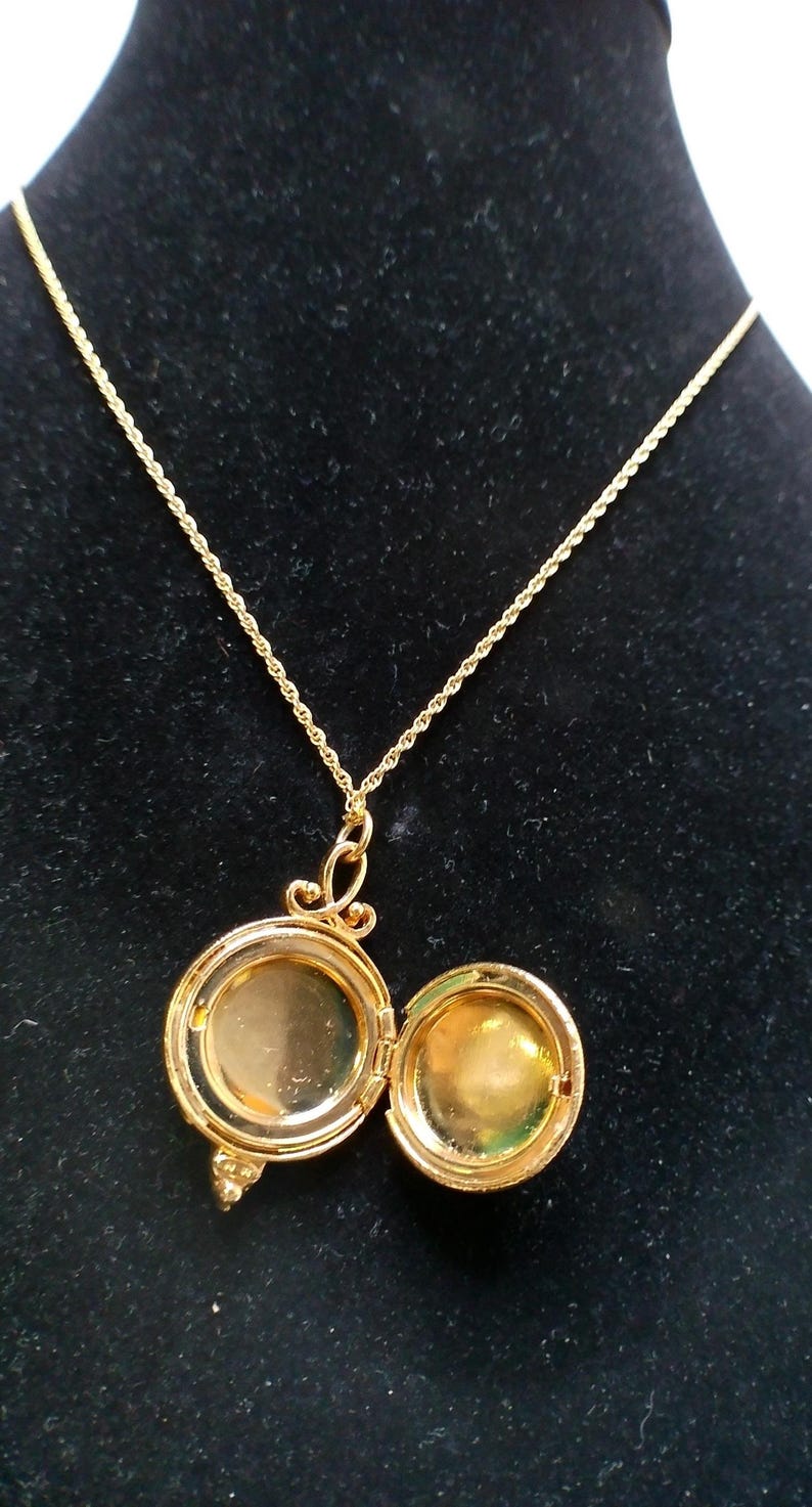 May include: A gold-colored locket necklace with an open, round pendant. The pendant is hinged and opens to reveal a small space. The necklace chain is a delicate, twisted design. The locket is displayed against a black background.