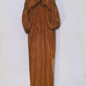 May include: A carved wooden figure of a religious person, with a golden halo. The figure is dressed in long robes and has a serene expression. The wood has a warm, brown tone, and the carving shows visible texture.