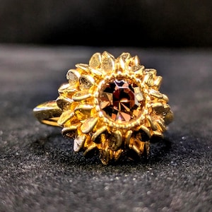 May include: A gold ring with a central brown gemstone, encircled by a floral design. The ring's band is slender and polished, and the setting has a detailed, textured appearance. The ring is set against a dark background.