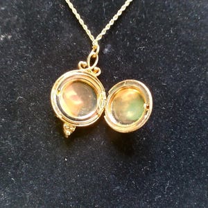 May include: Gold-toned locket necklace with an open, round pendant. The pendant is hinged and opens to reveal a small space. The chain is a delicate rope style. The background is a dark, solid color.