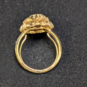 May include: A gold ring with a floral design on the face. The ring has a double band and a textured, detailed flower-like setting. The ring is set against a dark background.