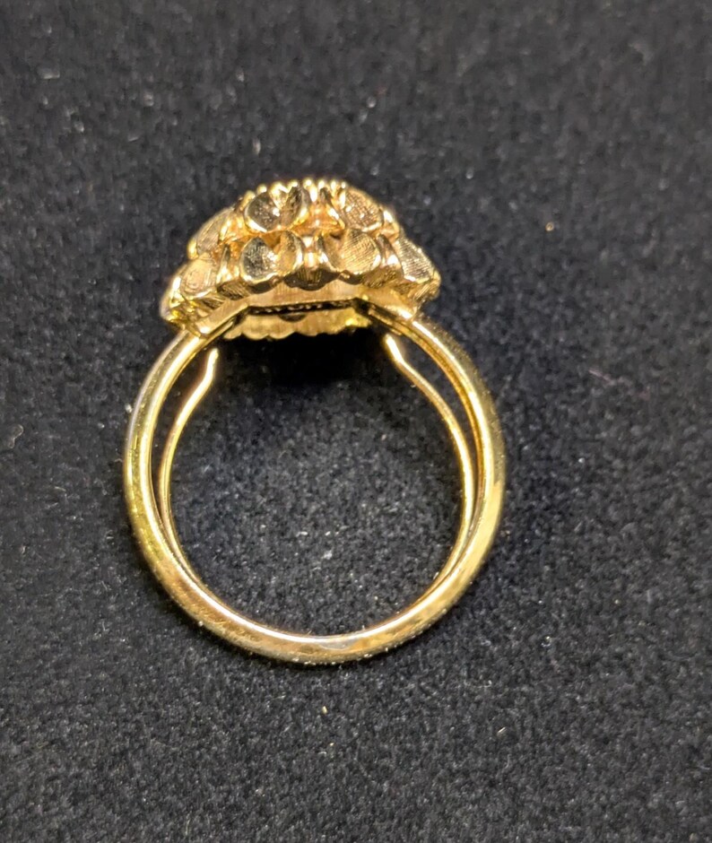 May include: A gold ring with a floral design on the face. The ring has a double band and a textured, detailed flower-like setting. The ring is set against a dark background.