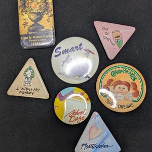 Puede incluir: Una colección de pines y botones vintage con varios diseños. Incluye un pin rectangular con un diseño floral y texto, un botón redondo "Smart Ass" y un pin triangular con un gráfico de momia.