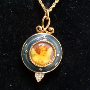 May include: Gold-toned necklace featuring a pendant with a painted cherub image. The pendant has a dark blue enamel border with small crystal accents and a heart-shaped cluster of crystals at the bottom. The chain is a delicate rope style.