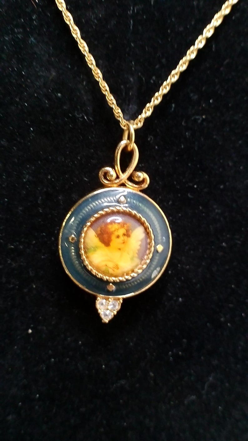 May include: Gold-toned necklace featuring a pendant with a painted cherub image. The pendant has a dark blue enamel border with small crystal accents and a heart-shaped cluster of crystals at the bottom. The chain is a delicate rope style.