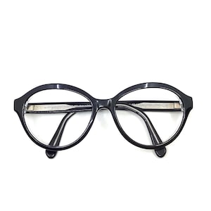 May include: Black oval-shaped eyeglasses with a silver accent on the frame. The glasses have a classic design with a thick frame and thin temples. The lenses are clear, and the glasses are set against a white background.
