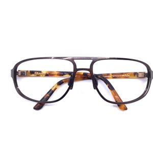 May include: Brown metal-framed eyeglasses with a double bridge and clear lenses. The temples feature a tortoiseshell pattern. The glasses are set against a white background.
