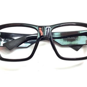 May include: Black-framed eyeglasses with a thick, rectangular design. The glasses have a glossy finish and feature a textured arm. The lenses are clear, and the frame has a modern, bold aesthetic. The text "MJ814-02" is visible on the arm.