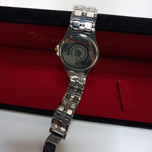May include: A silver-toned wristwatch with a stainless steel band, displayed in a red velvet box. The watch face is not visible, but the back shows the brand logo and model number. The band is partially open, revealing the clasp.