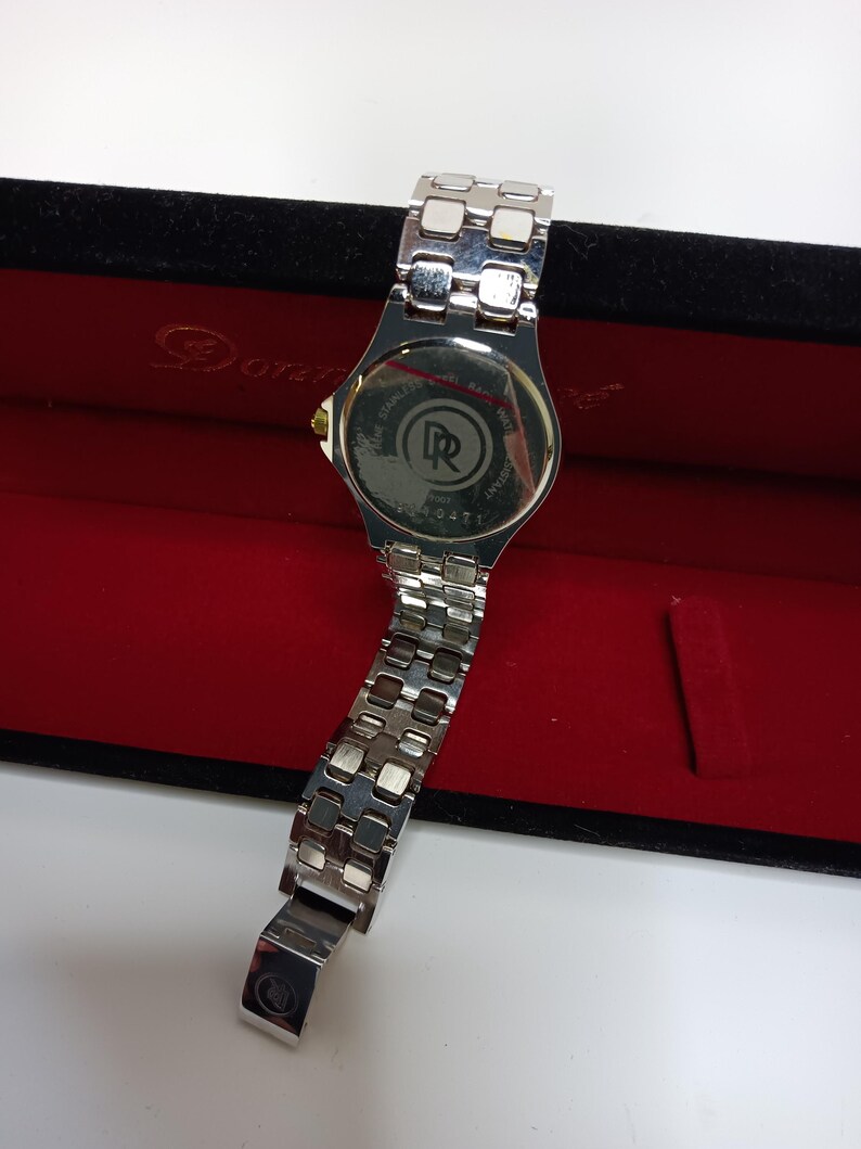 May include: A silver-toned wristwatch with a stainless steel band, displayed in a red velvet box. The watch face is not visible, but the back shows the brand logo and model number. The band is partially open, revealing the clasp.