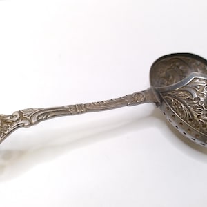 Pode incluir: Um coador de chá prateado antigo com um design floral ornamentado. O coador tem uma alça longa e detalhada e uma cabeça perfurada em forma de tigela. O design inclui padrões intrincados e pequenos orifícios para coar as folhas de chá. Um utensílio de cozinha vintage.