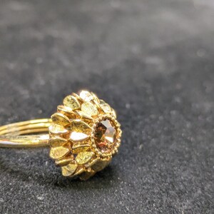 May include: A gold-colored ring with a floral design. The ring features a cluster of gold petals surrounding a central, brown gemstone. The band is thin and simple, and the ring is set against a dark background.
