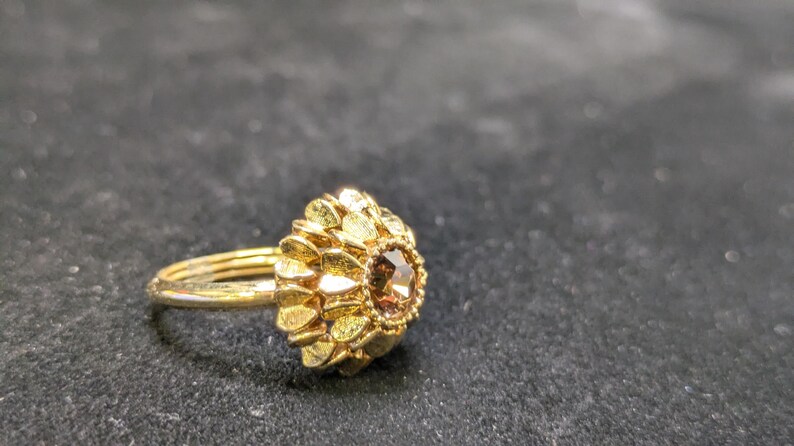 May include: A gold-colored ring with a floral design. The ring features a cluster of gold petals surrounding a central, brown gemstone. The band is thin and simple, and the ring is set against a dark background.
