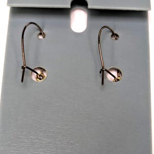 May include: A pair of earrings with a dark metal hook design, displayed on a gray card with a hole at the top. The earrings have a small, round, gold-colored accent near the hook.