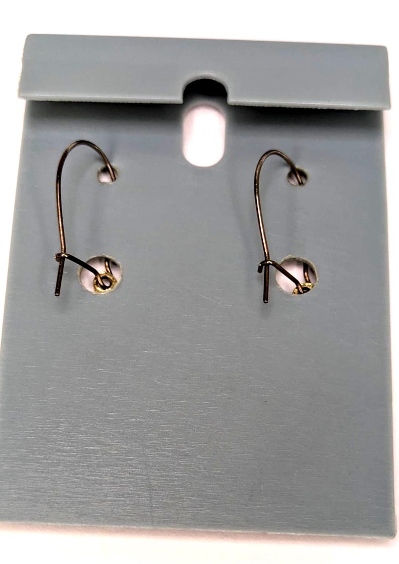 May include: A pair of earrings with a dark metal hook design, displayed on a gray card with a hole at the top. The earrings have a small, round, gold-colored accent near the hook.