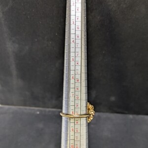 May include: A gold-colored ring with a floral design is placed on a ring sizer. The ring is measured at approximately a size 8. The ring sizer is silver with black markings and numbers.