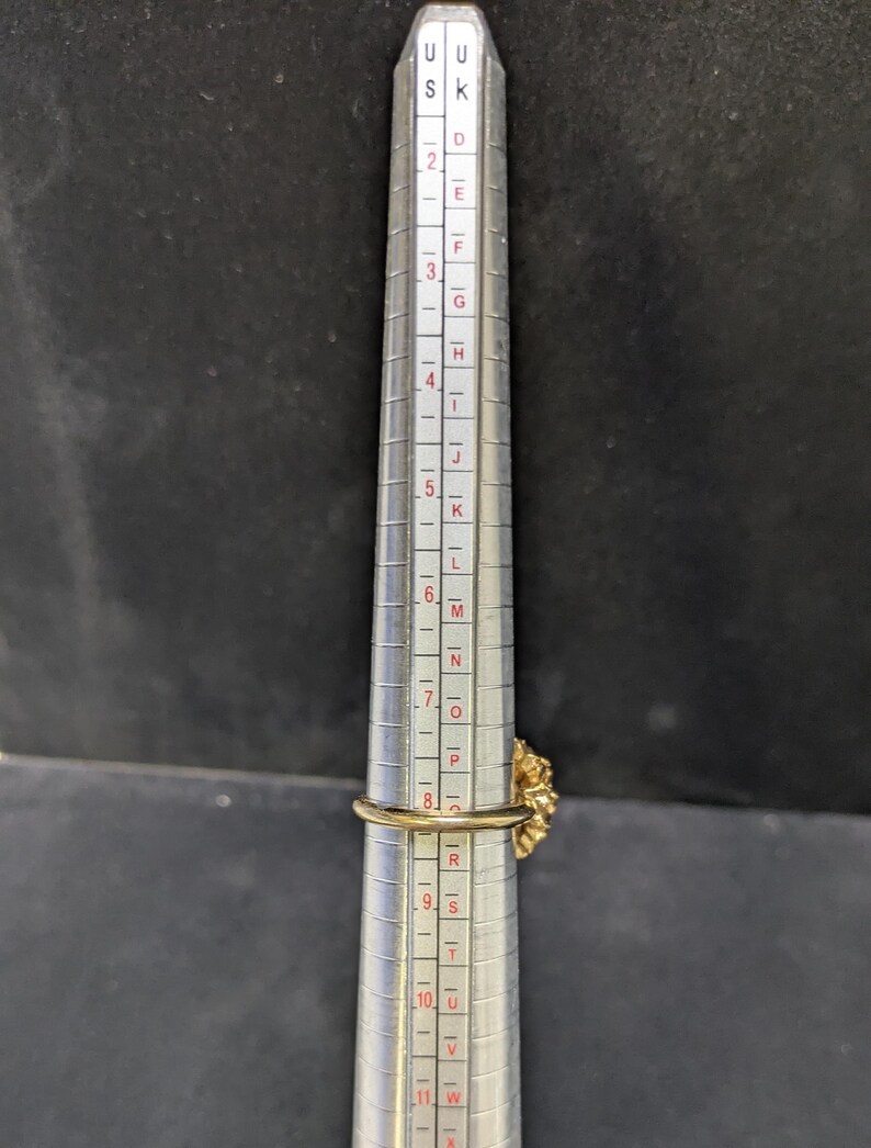 May include: A gold-colored ring with a floral design is placed on a ring sizer. The ring is measured at approximately a size 8. The ring sizer is silver with black markings and numbers.