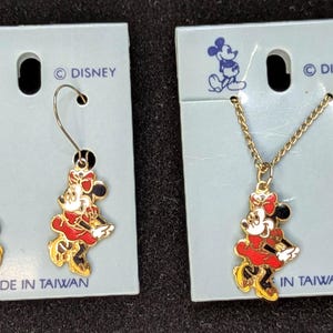 May include: A set of Disney Minnie Mouse jewelry, including a pair of earrings and a necklace. The earrings and pendant feature a red, white, and black Minnie Mouse design with gold-colored accents. The jewelry is attached to light blue cardboard packaging. The text "MADE IN TAIWAN" is visible.