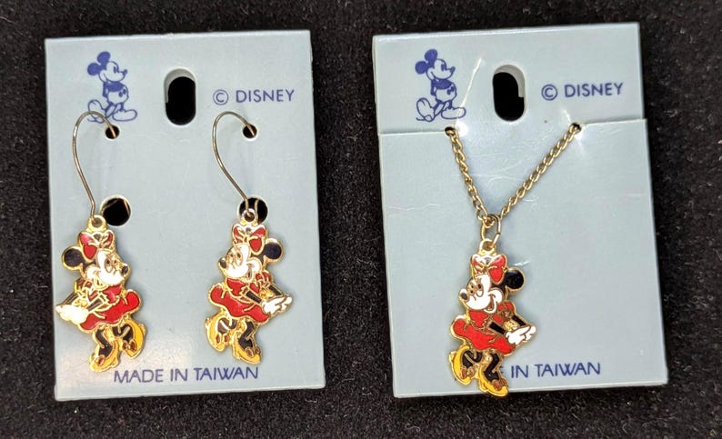 May include: A set of Disney Minnie Mouse jewelry, including a pair of earrings and a necklace. The earrings and pendant feature a red, white, and black Minnie Mouse design with gold-colored accents. The jewelry is attached to light blue cardboard packaging. The text "MADE IN TAIWAN" is visible.