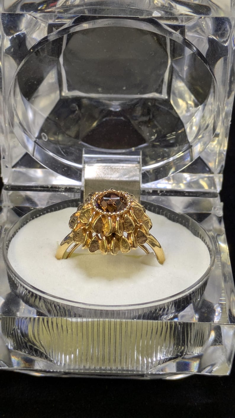 May include: A gold ring with a large, round, brown gemstone at the center, surrounded by smaller, petal-shaped gold accents. The ring is displayed in a clear, faceted jewelry box with a white interior.