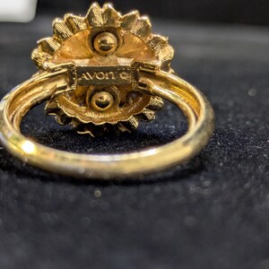 May include: A gold-toned ring with a floral design. The ring features a textured, flower-shaped setting with a central design. The band is smooth and rounded. The word "AVON" is stamped on the ring.