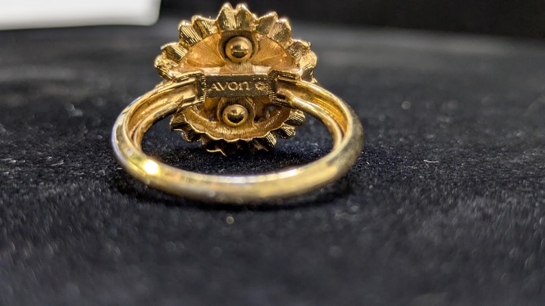 May include: A gold-toned ring with a floral design. The ring features a textured, flower-shaped setting with a central design. The band is smooth and rounded. The word "AVON" is stamped on the ring.