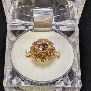 May include: A gold ring with a central, round, light-colored stone, surrounded by a detailed, flower-like setting. The ring is displayed in a clear, hinged, plastic box with a white interior. The box is on a dark surface.