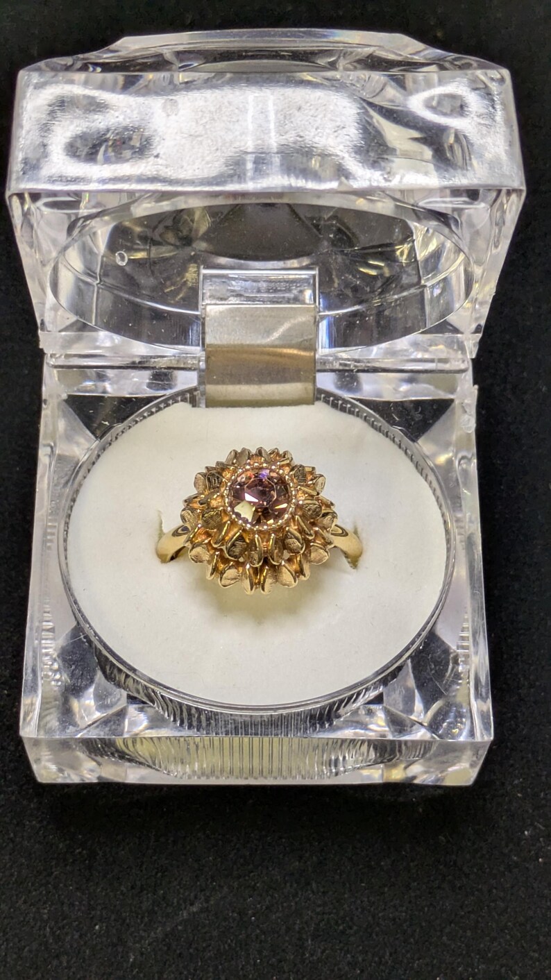May include: A gold ring with a central, round, light-colored stone, surrounded by a detailed, flower-like setting. The ring is displayed in a clear, hinged, plastic box with a white interior. The box is on a dark surface.
