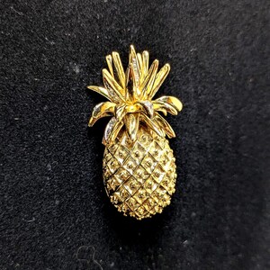 May include: A gold-coloured pineapple brooch. The brooch has a detailed, textured surface and a spiky crown. The pineapple shape is a classic design, and the gold colour gives it a luxurious look. The brooch is likely made of metal.