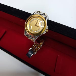 May include: A gold and silver wristwatch with a gold face and diamond accents. The watch is in a black box with a red interior. The watch band is silver and gold.