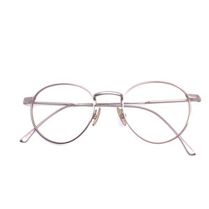 May include: Silver-toned, round-framed eyeglasses. The glasses feature a thin metal frame with a double bridge and curved temples. The lenses are clear, and the overall design is minimalist and modern. The glasses are set against a white background.