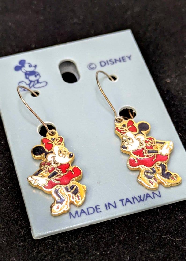 May include: A pair of gold-toned Minnie Mouse dangle earrings. Each earring features a red dress, black ears, and a white face. The earrings are on a light blue card with the Disney logo and "MADE IN TAIWAN".