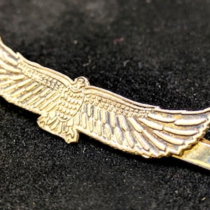 May include: A gold-colored metal hair clip in the shape of an eagle with outstretched wings. The clip is detailed with feather patterns and has a rectangular clasp. The clip is on a black surface.