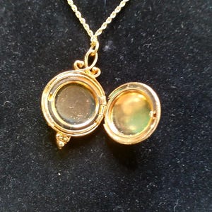 May include: A gold-colored locket necklace with an open, round pendant. The pendant is hinged and opens to reveal a small space. The necklace chain is also gold-colored and has a twisted rope design. The locket is on a black background.