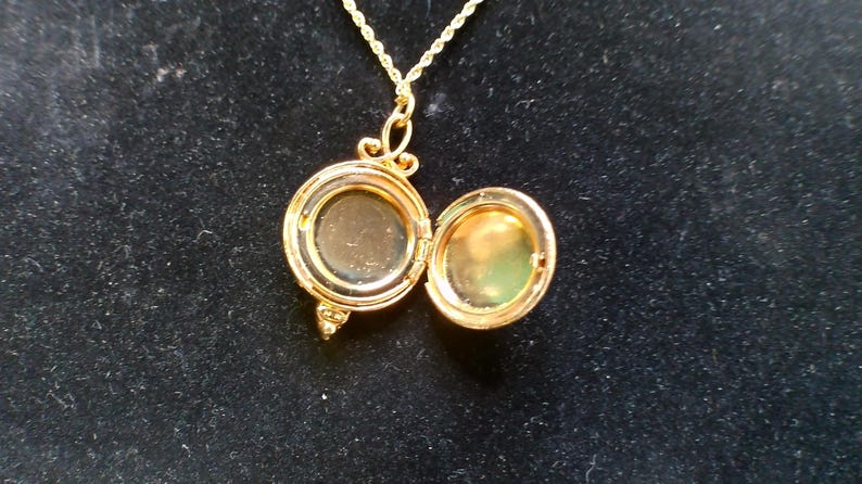May include: A gold-colored locket necklace with an open, round pendant. The pendant is hinged and opens to reveal a small space. The necklace chain is also gold-colored and has a twisted rope design. The locket is on a black background.