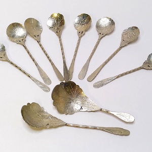 Op de afbeelding: Een verzameling zilverkleurige lepels met sierlijke bloemmotieven op de lepels. De lepels hebben gedraaide handvatten en zijn in een waaier op een wit oppervlak gerangschikt. De lepels zijn waarschijnlijk om te serveren of te eten.