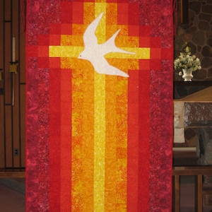 May include: A red and orange quilted banner with a white dove flying over a yellow cross. The banner has a fringed border.