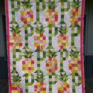 Puede incluir: Una colcha con un diseño de patchwork que presenta piñas en tonos de amarillo, rojo y verde. La colcha está hecha con una variedad de telas y tiene un borde rosa.