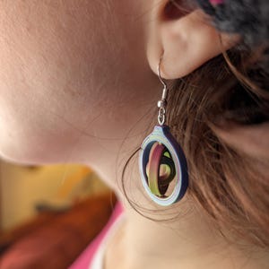 Könnte beinhalten: Nahaufnahme eines Ohrrings aus blauem, grünem und rosa Papier. Der Ohrring hat ein kreisförmiges Design mit einem silbernen Haken. Das Papier ist spiralförmig angeordnet und schafft ein einzigartiges und farbenfrohes Accessoire.