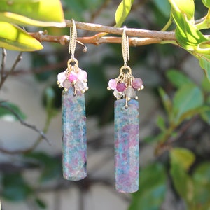 May include: A pair of dangle earrings featuring long, rectangular, mottled teal and magenta stone pendants. Each earring is topped with a cluster of small, faceted pink, red, and translucent grey beads attached to gold-toned metal ear wires.