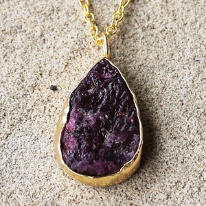 May include: A gold chain necklace with a teardrop-shaped pendant featuring a raw, purple ruby gemstone.