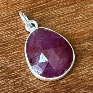 May include: A teardrop-shaped pendant featuring a faceted, deep magenta-coloured gemstone set in a simple silver-toned metal bezel. The pendant includes a small circular silver-toned jump ring at the top for attachment to a chain or cord.