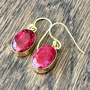 May include: A pair of gold drop earrings with oval, deep red gemstones. The gemstones are set in a gold bezel, and the earrings have a simple hook closure. The earrings are displayed on a wooden surface.