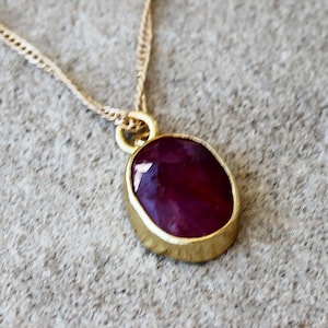 Może przedstawiać: Złoty naszyjnik z wisiorkiem z dużą, owalną, ciemnoczerwoną kamieniem szlachetnym. Kamień jest osadzony w złotej oprawie i zawieszony na delikatnym złotym łańcuszku. Nieregularny kształt wisiorka dodaje mu wyjątkowego uroku.