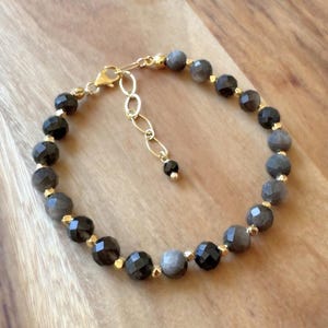 May include: A beaded bracelet featuring alternating faceted dark grey and black round beads separated by small gold-toned metal spacers. The bracelet includes a gold-toned lobster claw clasp and an adjustable chain extension with a single bead.